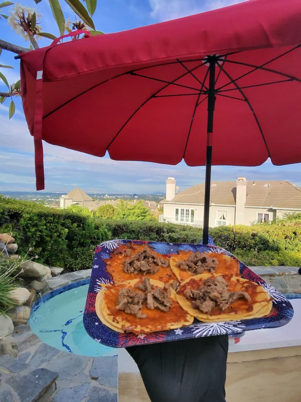 Four beef tacos on a tray under a red umbrella overlooking a scenic hillside.