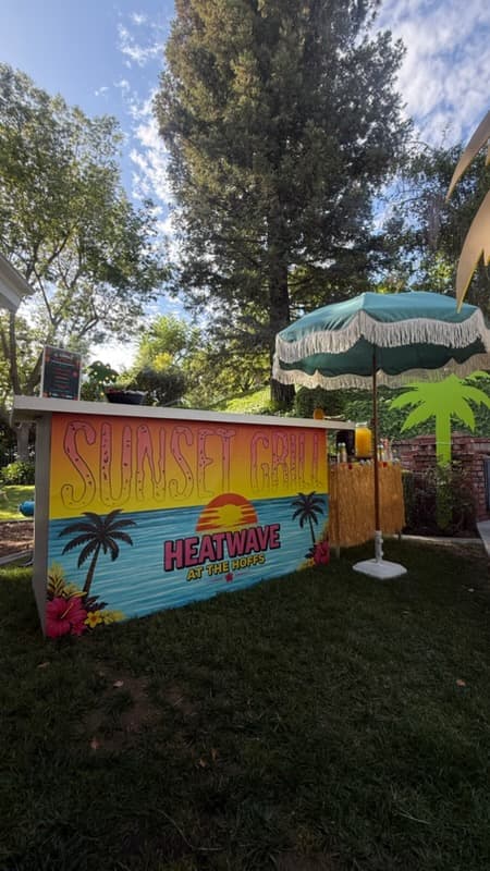 Tropical backyard bar with Sunset Grill graphics and a green fringed umbrella on grass.