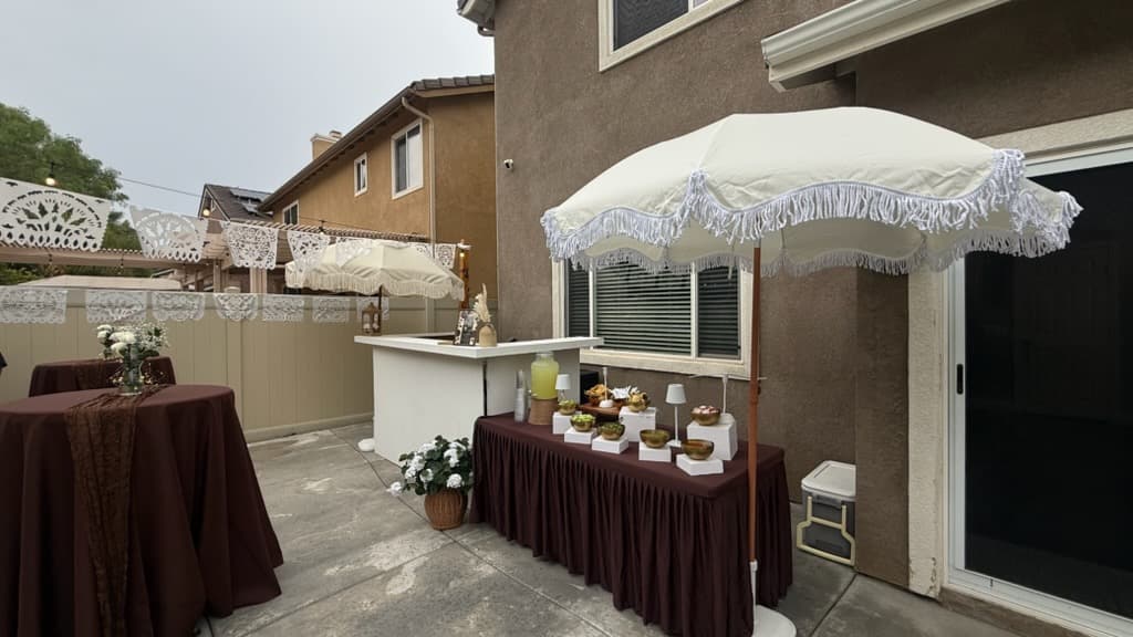 Outdoor party setup featuring brown-clothed tables, white fringed umbrellas, and decorative white paper banners.