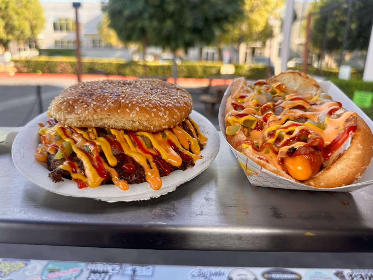 Sesame seed burger and loaded hot dog, both drizzled with mustard and orange sauces.