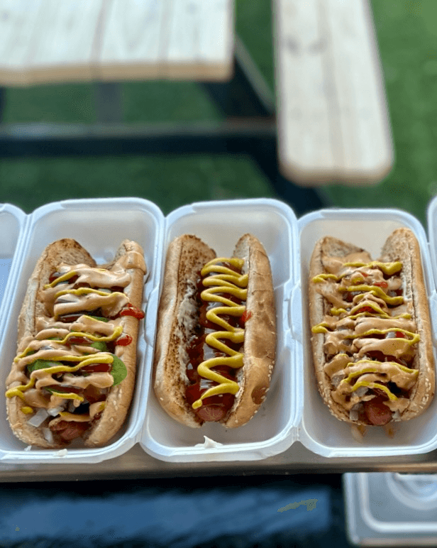 Three toasted hot dogs topped with mustard, ketchup, and creamy sauce in white containers outdoors.